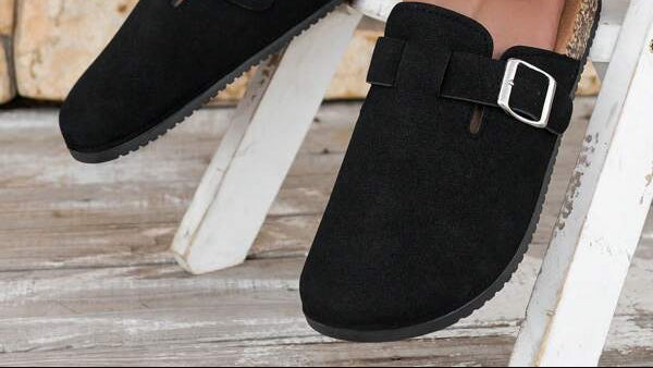 Black suede clogs with a buckle worn by a person sitting on a stone wall.